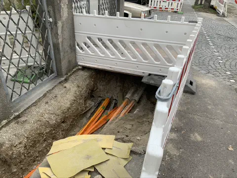 Aufgegrabener Gehweg mit orangefarbenen Glasfaser-Leerrohren hinter Absperrung